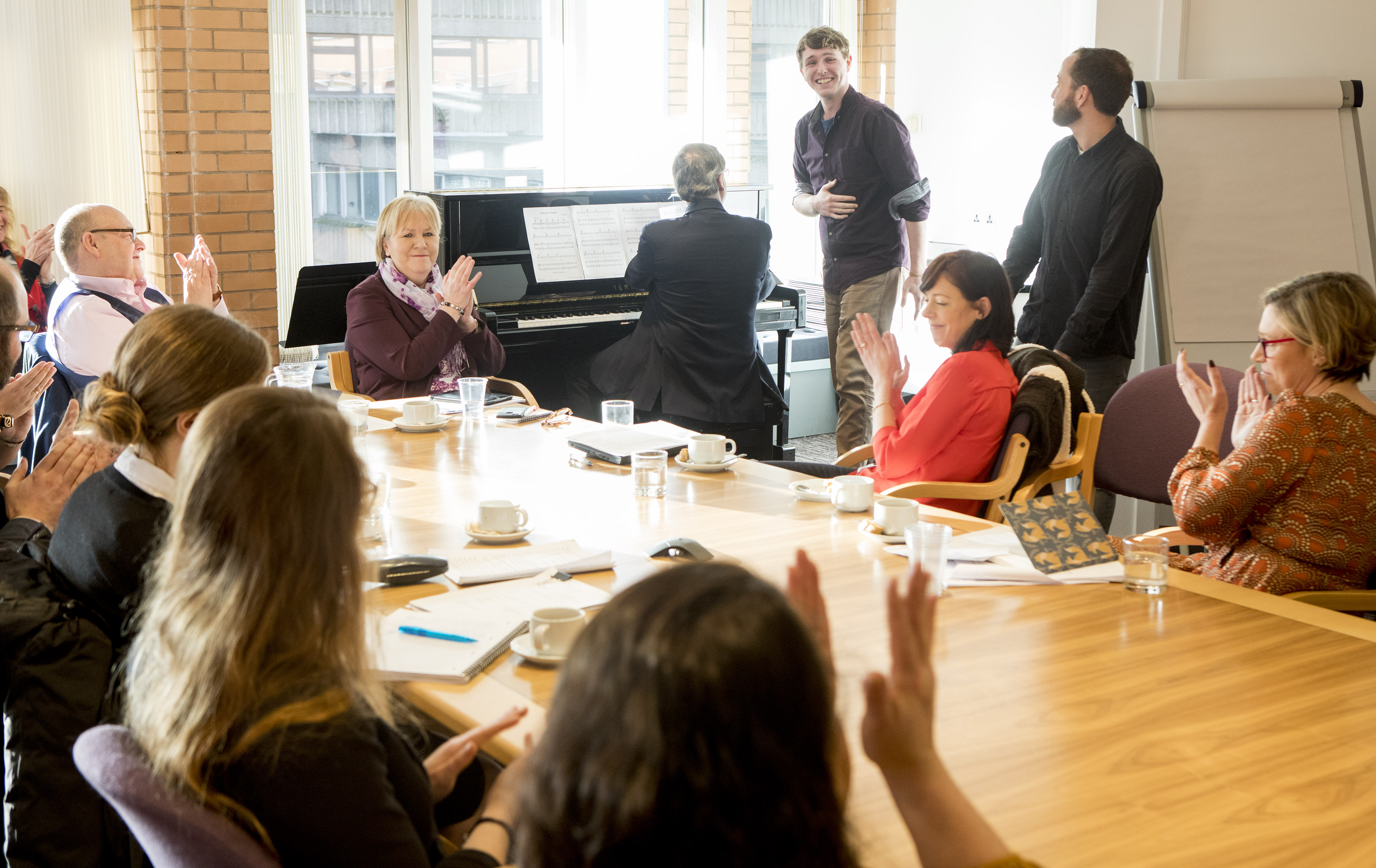 Photograph of Committee Members and students at the Royal Conservatoire of Scotland around a table long table, clapping as a musician plays the piano.